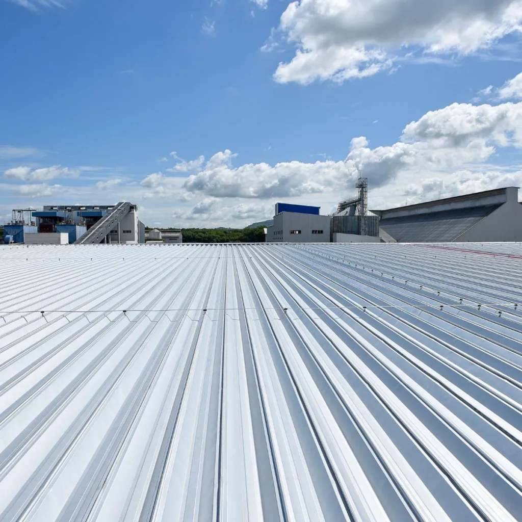 A wide, white metal rooftop stretches into the distance under a partly cloudy blue sky, with industrial buildings and structures visible in the background.