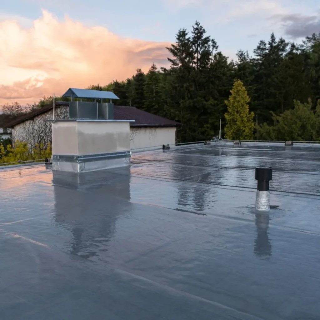 A flat roof covered with a shiny waterproof membrane, featuring a vent and a chimney stack. Trees and a building are visible in the background under a cloudy sky at sunset.