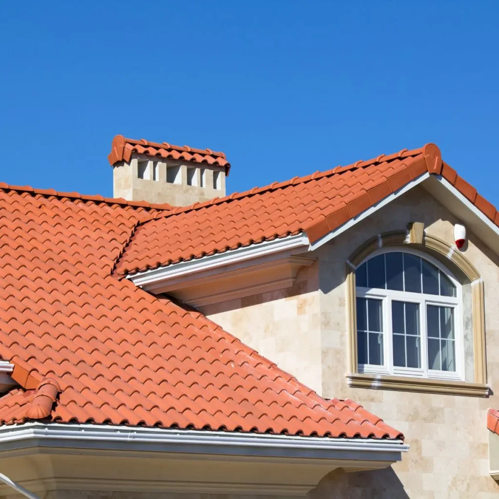 A house with beige stone walls and a red tiled roof, featuring a white-framed arched window and a chimney, under a clear blue sky.
