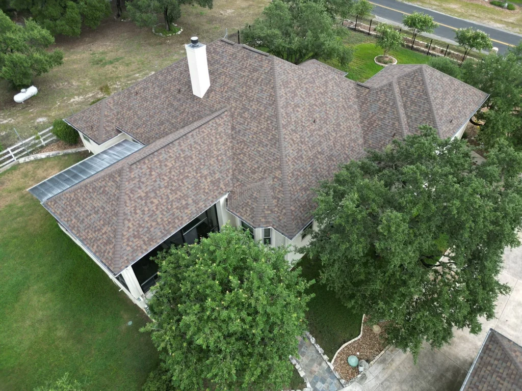 Aerial view of a house with a complex, brown shingle roof surrounded by trees, a grassy yard, a driveway, and a nearby road. A chimney and a screened patio are visible on the house.