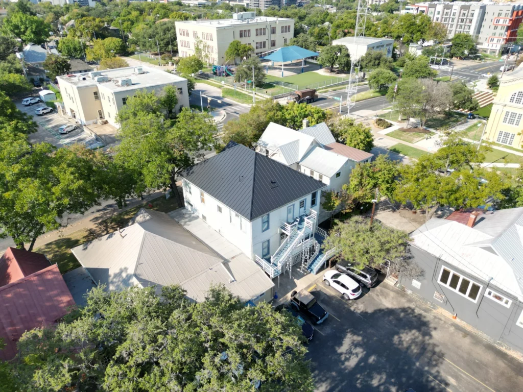 Aerial view of a neighborhood with several buildings, trees, parked cars, and a few open spaces visible on a sunny day. A white building with a metal staircase stands prominently in the center.
