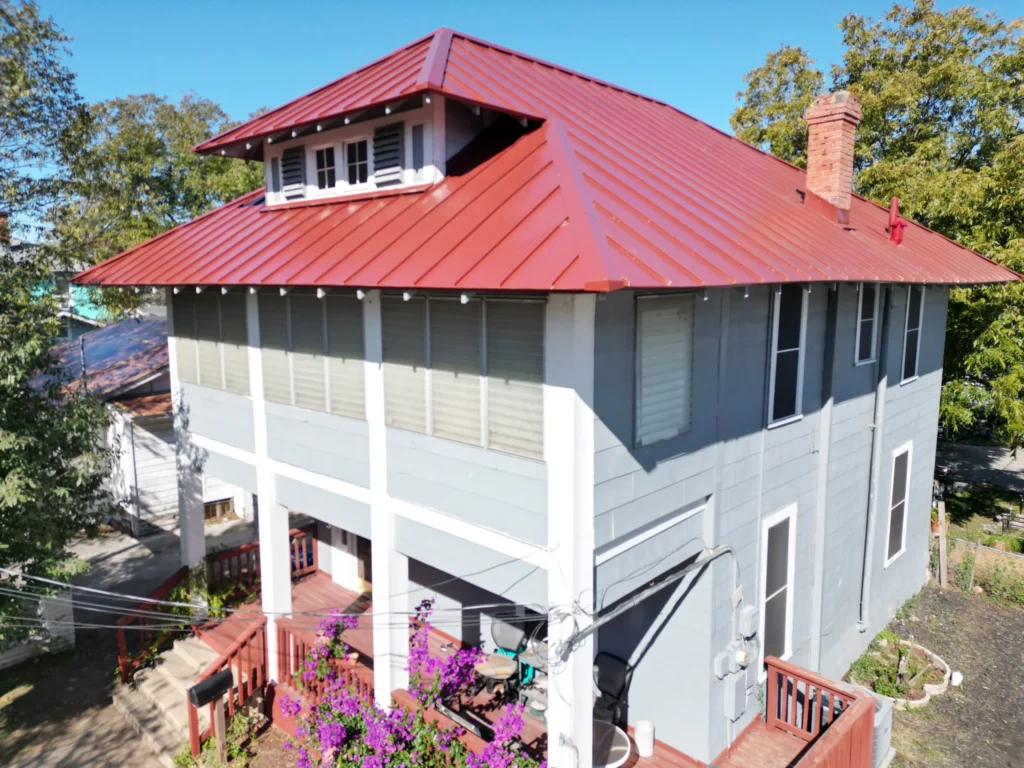 A two-story gray house with a red metal roof, white trim, and a small upper dormer window. There are multiple windows, a brick chimney, and a porch with purple flowers and greenery in the front yard.