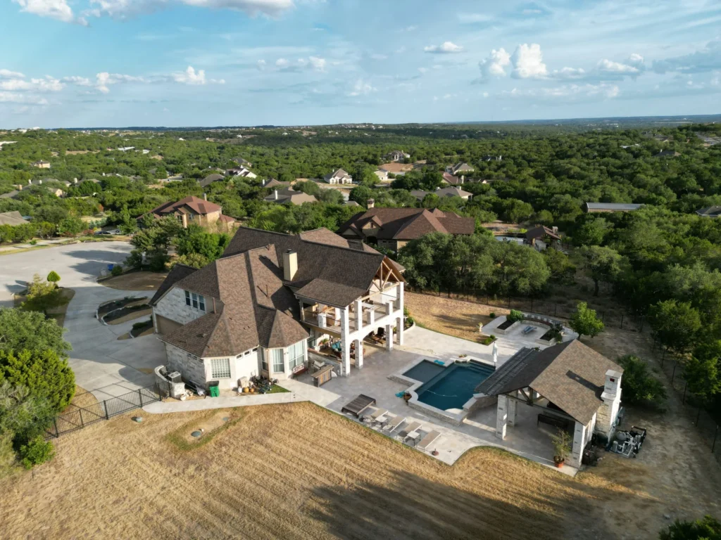 Aerial view of a large, two-story house with a brown roof, spacious patio, and swimming pool, surrounded by trees, other homes, and a wide open landscape under a partly cloudy sky.