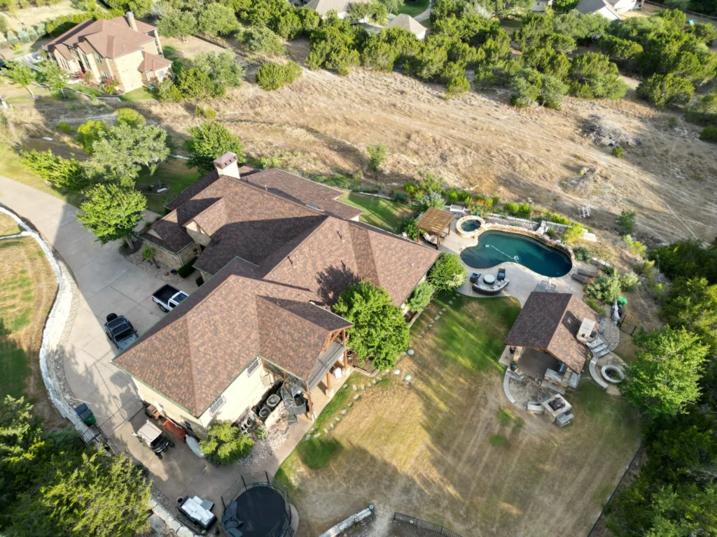 A large house with a brown roof sits on a spacious lot, surrounded by trees. The backyard features a swimming pool, hot tub, and seating area. A driveway and parked vehicles are visible. The area appears suburban and green.