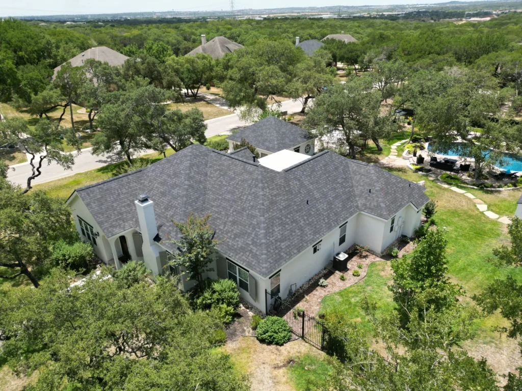 Aerial view of a white, single-story house with a dark gray roof, surrounded by trees and greenery; neighboring homes and a swimming pool are visible in the background.