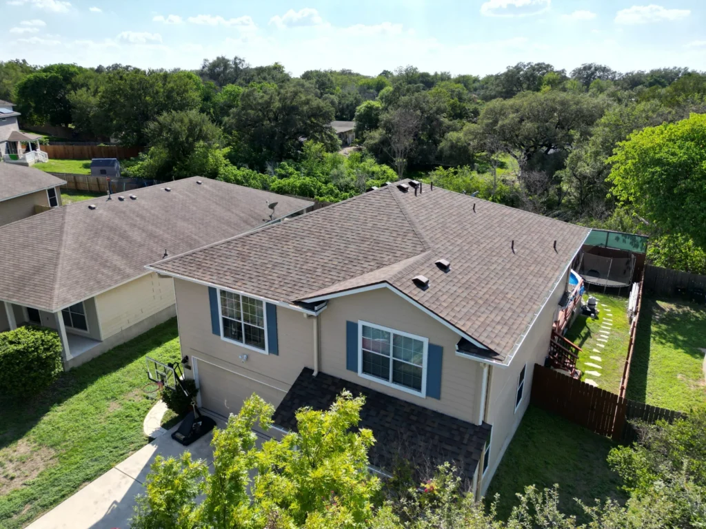 A two-story beige house with brown shingles, surrounded by green trees and grass, sits in a suburban neighborhood on a sunny day. A fenced backyard with a small area for outdoor activities is visible.