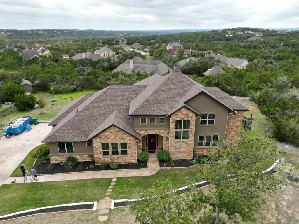 A large two-story house with stone and tan siding stands amid a green, hilly neighborhood. There is a driveway, lawn, and above-ground pool on the left, with trees and other homes in the background.