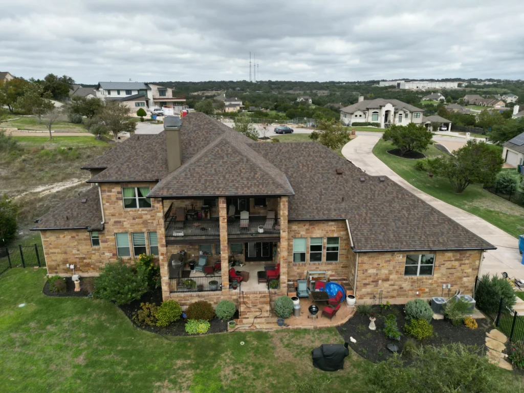 Aerial view of a large stone house with two covered patios, outdoor furniture, and a neatly landscaped backyard in a suburban neighborhood under a cloudy sky.