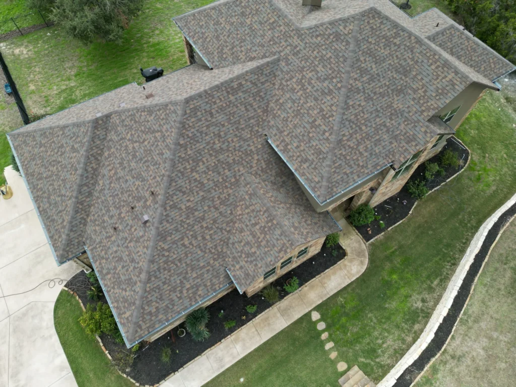 Aerial view of a house with a complex, multi-gabled brown shingle roof, stone exterior walls, and landscaped yard with a curved walkway and green lawn.