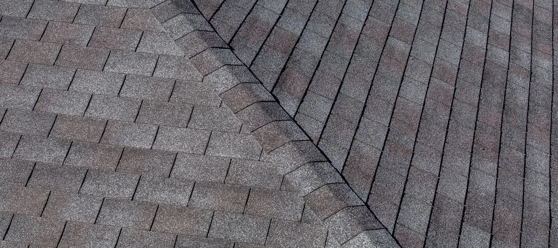 Close-up view of an asphalt shingle roof, showing the intersection of two sloped sections with overlapping gray shingles. The pattern highlights the ridge where the two roof planes meet.