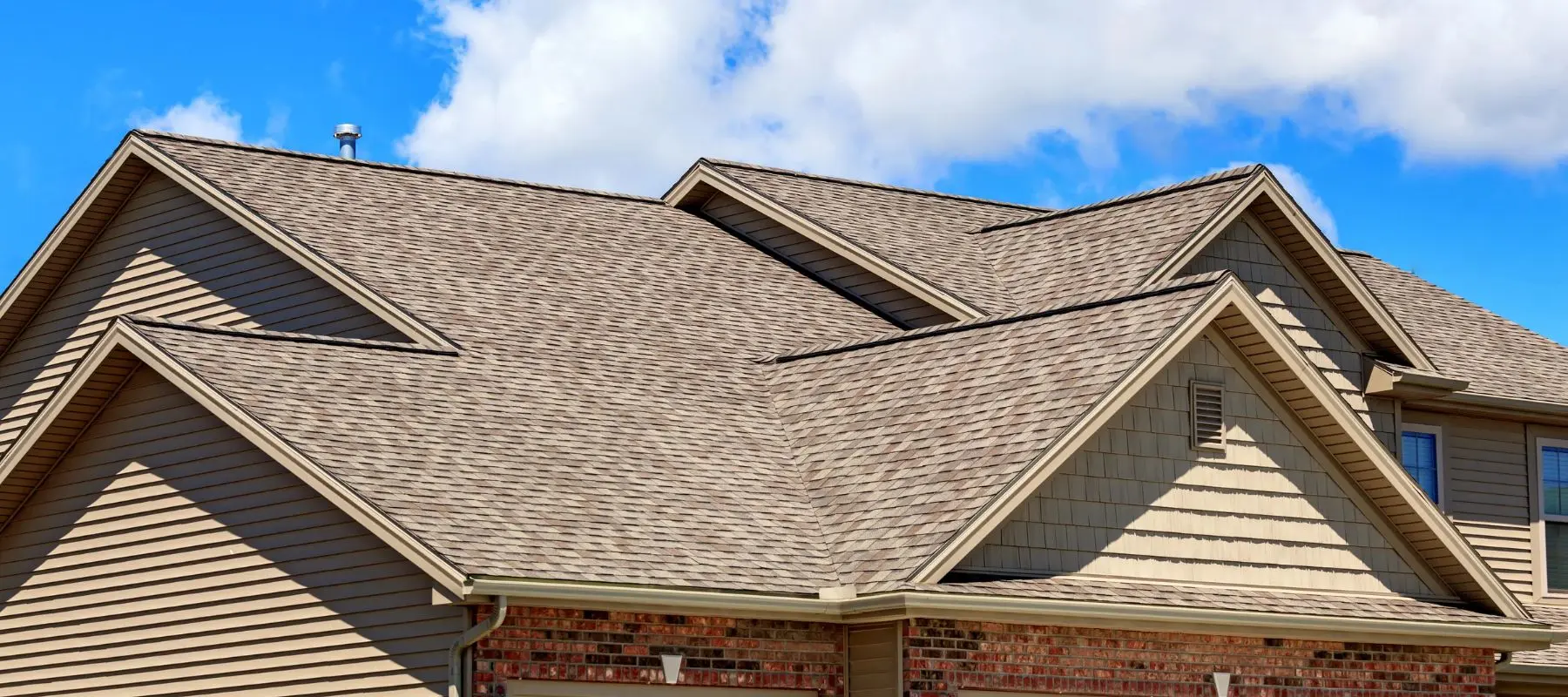 A multi-gabled house roof covered in brown asphalt shingles against a blue, cloudy sky.